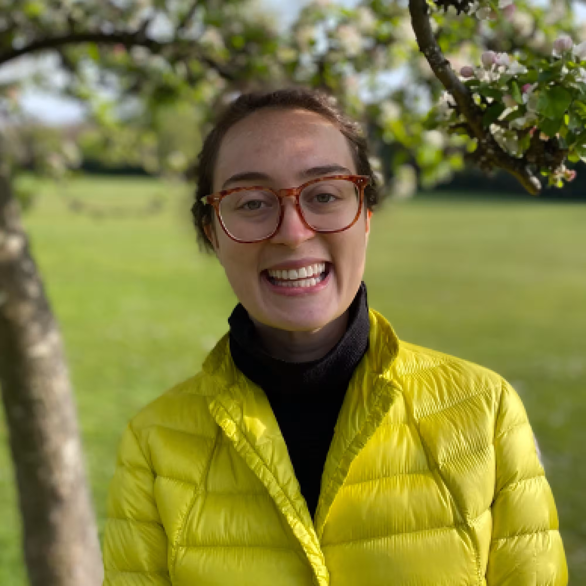 Portrait of Jessica Renzella, a young lady with brown hair and glasses, standing under a tree by a field.