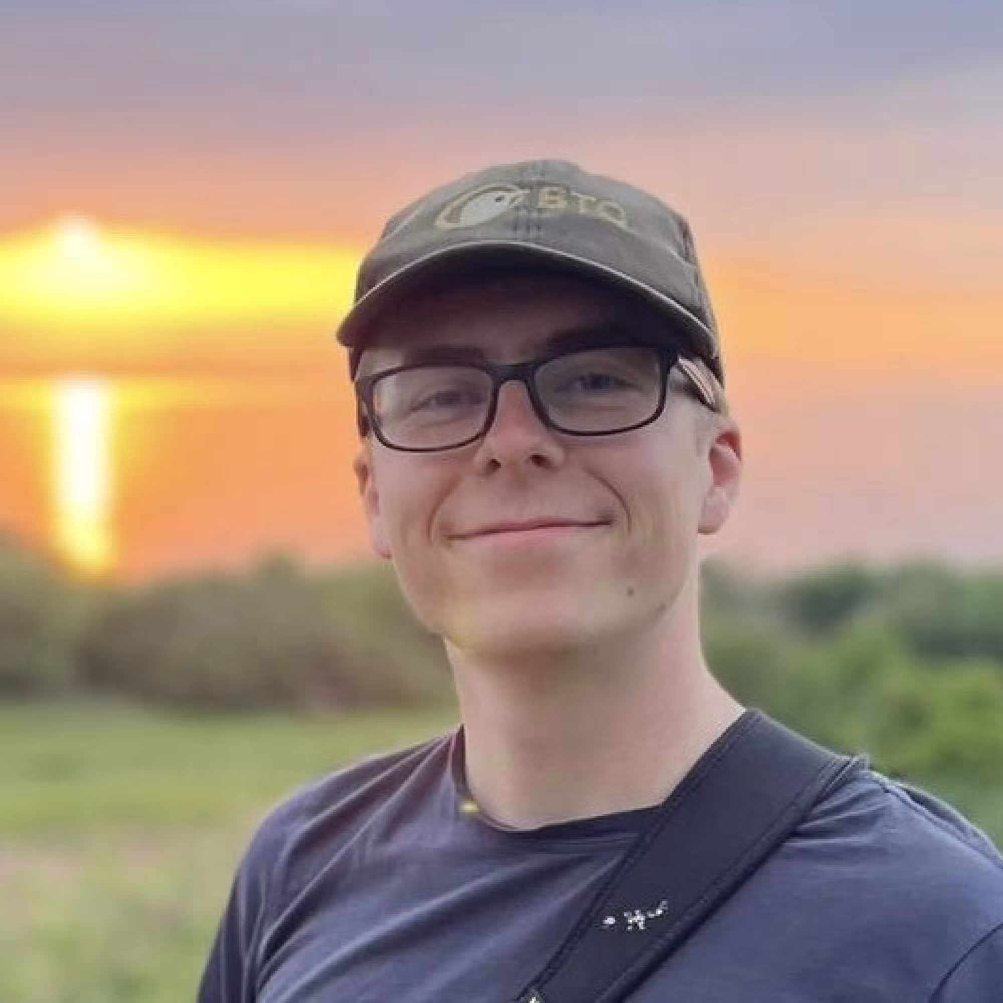 Portrait of Patrick Lewin, a young white man standing in a field at sunset.