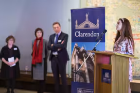 A woman stands at a podium with the Clarendon banner in the background, with 3 people watching