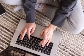 A person sits cross-legged on a carpet using a laptop