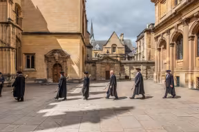 Men and women in black University robes processing between two buildings