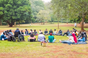 Group of people sitting in a circle on grass in a park in Oxford during an outdoor community discussion, including a participant using a wheelchair