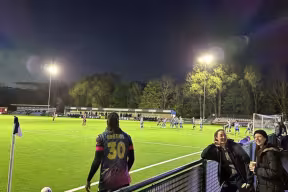 Evening football match at a floodlit stadium, with players on the pitch, a player wearing number 30 near the touchline, and spectators watching from the stands along the sideline.