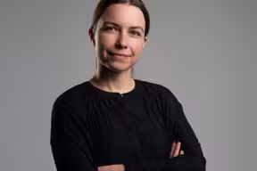 Portrait of a person wearing a black long-sleeved top, standing with arms crossed against a plain grey background.