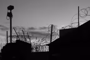 Security camera mounted on a wall covered in razor wire in silhouette in monochrome