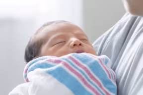 An infant wrapped in a striped blanket resting in the arms of an adult in a softly lit indoor setting.
