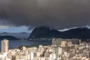 Wide cityscape with dense white and beige buildings along a coastline, backed by dark mountains and islands across the water, under heavy, low storm clouds.