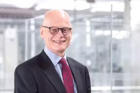 A person wearing glasses, a dark suit, light blue shirt, and red patterned tie, standing indoors in a bright, modern office with glass walls in the background
