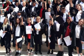 A large group of students wearing graduation gowns, standing on steps outdoors and holding certificates, with some raising their caps.
