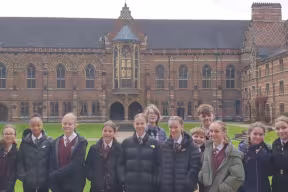 A group of school students in uniform standing on a lawn in front of a red-brick university college building with arched windows and a central entrance.