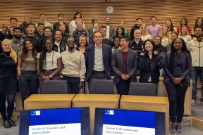  Large group of people standing and seated in a tiered lecture theatre, posing for a group photo, with curved wooden wall panels behind them and presentation screens visible at the front.