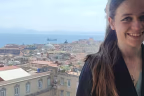 Person standing near a window or balcony, photographed from the waist up, with a coastal city, sea, and distant mountains visible in the background.