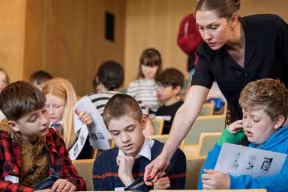 Students seated in a tiered lecture room reading worksheets, while an adult leans over to assist one student, with other students visible in the background.