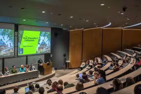 Wide view of a modern lecture theatre with tiered seating filled with attendees, a panel and speaker at the front, and two large presentation screens displaying a Vice‑Chancellor’s Colloquium slide.