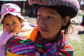A young adult carrying a baby in a woven sling outdoors, with both wearing colorful clothing in a rural setting.
