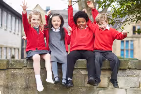 Four smiling primary school children in red school uniforms sitting on a stone wall outdoors with their hands raised.