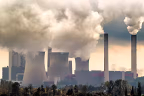 Chimneys of a coal power plant release fumes of pollution into the sky. In the foreground is a line of trees.