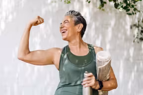 Smiling older woman in green activewear flexing her arm outdoors while holding a yoga mat and water bottle.