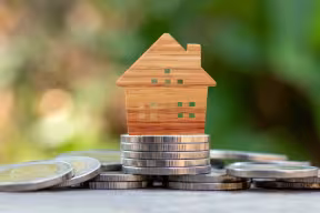 Wooden house model placed on stacked coins with blurred greenery in the background, representing housing and finance.