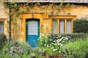Traditional honey-coloured stone cottage with a blue door surrounded by flowers and greenery.
