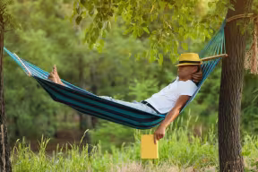 Person relaxing in a hammock between trees outdoors while holding a book.
