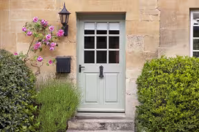 Front door of a stone house with a pale green frame, surrounded by leafy shrubs and pink climbing roses.