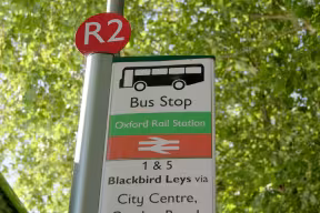 Bus stop sign displaying routes towards Oxford city centre, against a background of foliage.