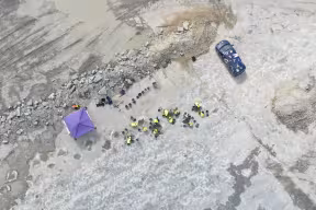 Drone shot over a quarry with a line of dinosaur footprints, a team of people in helmets and high-vis jackets excavating them, a gazebo and a minibus