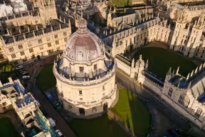 An aerial shot over the Radcliffe Camera (a circular building with a dome) and All Souls College (historic building with a grassy quad)