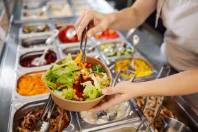 Close up of a person serving themselves a bowl of plant-based salad (lettice, chickpeas, peppers, beetroot) at a canteen.