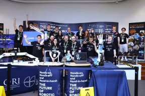 A diverse group of men and women wearing identical T Shirts wave enthusaistically behind Oxford Robotics Institute's exhibition stand featuring robotic arms and soft robotic hands.