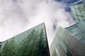 Upward-looking view of several modern glass office buildings with sharp, angular architecture. The reflective façades mirror tall green trees, creating the illusion of a forest inside the buildings. A cloudy sky fills most of the frame, with patches of blue visible between the clouds, emphasizing the contrast between urban design and nature
