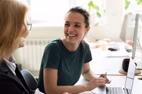 Two people smiling and talking in front of laptop in work setting