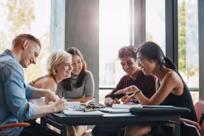 Five students working together around a table, discussing notes and study materials