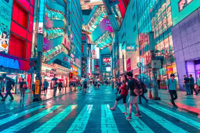 A vibrant city street at night lined with brightly lit shops, digital billboards, and illuminated buildings. People walk across a pedestrian crossing and along wet pavements reflecting colourful neon lights, creating a busy urban atmosphere. Elevated walkways and Japanese signs suggest a modern commercial district in Japan.