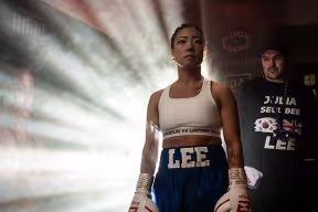 Julia Lee stands in a boxing ring wearing gloves and blue shorts, illuminated by dramatic light beams behind her as she prepares to compete, with a coach standing nearby