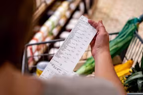 Close-up over-the-shoulder shot of a woman looking at a receipt after shopping at the supermarket, with a shopping basket of food in the background.