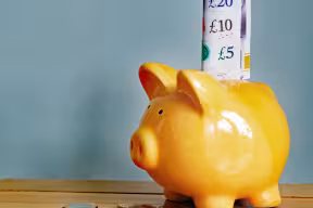 A yellow piggy bank with a £20, £10 and £5 note sticking out of the top sits on a table with coins scattered around it.