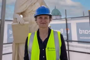 The Vice-Chancellor wears a blue hard hat on some scaffolding, with a muse statue behind her, and looks at the camera.  