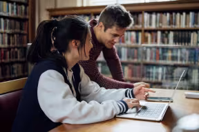 Two students working at a laptop in a library. 