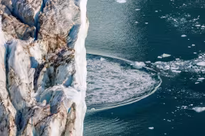 The front edge of a glacier above a fjord in Greenland. The ripples of a giant wave can be seen spreading across the water.
