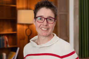 A portrait photo of Charlotte Deane, a white lady with short brown hair wearing glasses and a stiped jumper, standing by a bookcase.