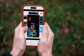 A person's hands hold a mobile phone with the screen facing towards the camera. The phone shows the video game Tetris, seen as a grid of different coloured squares against a black background.