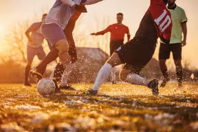 A group of male soccer players of mixed nationalities playing a match on a soccer field on winter day outdoors. Two players are performing a tackle.