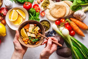 A spread of plant-based foods including flat breads, tomatoes, spring onions, oranges, carrots, blueberries, and garlic. In the foreground, two hands hold a bowl of lentil curry with tofu. 