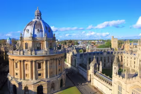 A view looking down over a large dome-shaped building. In the background is an Oxford college; a collection of buildings arranged in four sides around a quadrangle.
