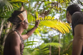 An Indigenous Brazilian young man from Guarani ethnicity in native dress shows the rainforest to a male tourist.