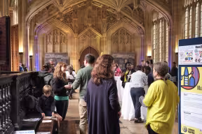 Groups of people look at posters displayed in a large historic stone hall. Light filters into the room through large windows. 
