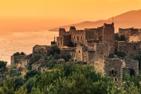 Old abandoned tower houses in Vathia Mani Peninsula at evening time, with the sun setting over the ocean. 