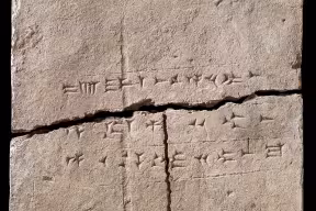 A clay tablet with wedge shaped marks (cuneiform script). The tablet is cracked horizontally and vertically into three pieces.